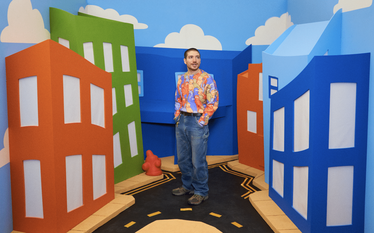 The artist, Danny Cole, stands in front of cartoon buildings while wearing a shirt with artwork on it.